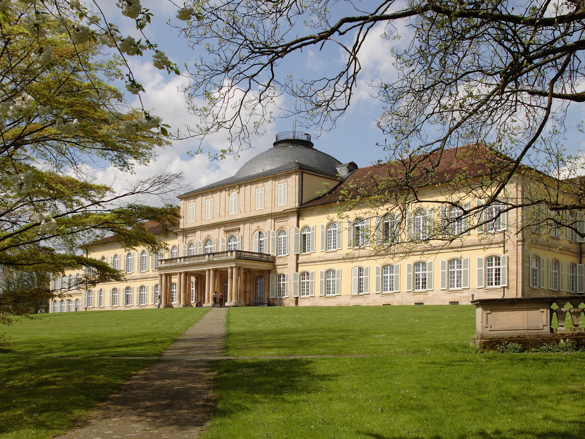 Das Bild zeigt das historische Schloss Hohenheim - ein sandfarbenes, prunkvolles Gebäude mit Balkon und großen Fenstern. Dieses ist eingebettet in die grüne Umgebung. Im Hintergrund blauer Himmel mit wenigen Wolken.